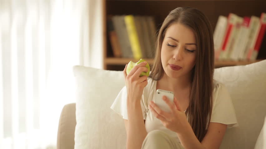 Pretty girl sitting on sofa using mobile phone eating apple and smiling