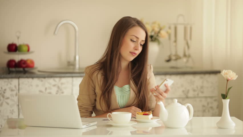 Pretty young woman sitting at kitchen table using mobile phone pouring tea into