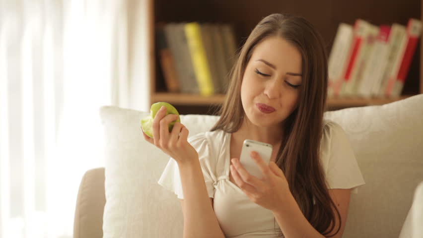 Pretty girl sitting on sofa eating apple using cellphone and smiling at camera