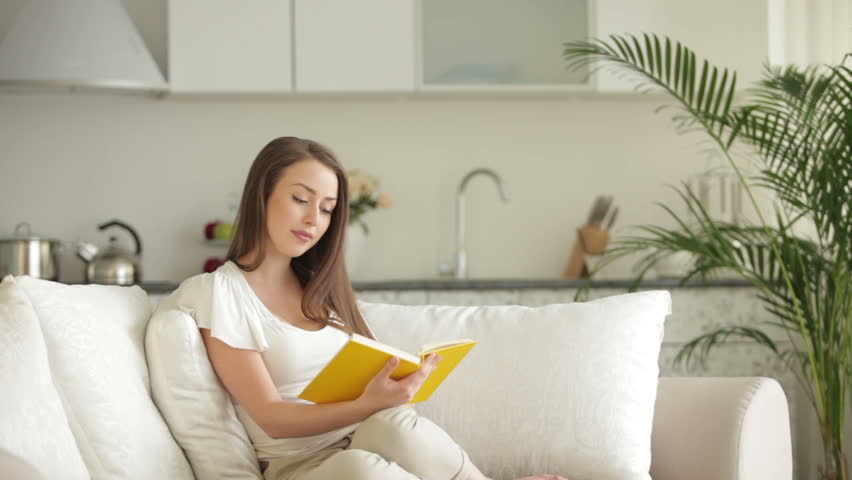 Pretty young woman relaxing on sofa with book and smiling at camera