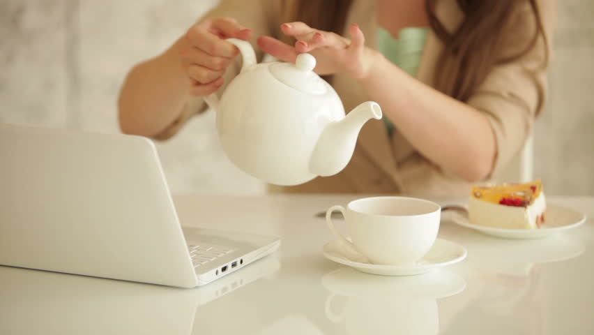 Young woman sitting at table with laptop pouring tea from teapot into cup