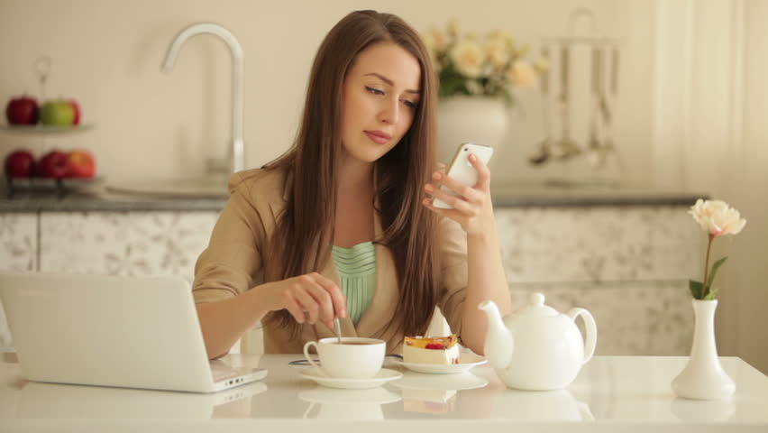 Pretty girl sitting at kitchen table with cup of tea using mobile phone and