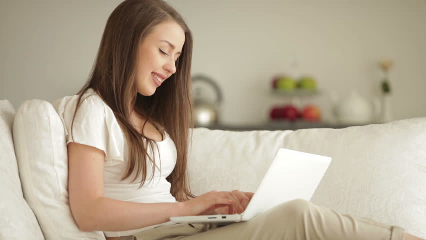Pretty girl sitting on sofa using laptop and smiling at camera