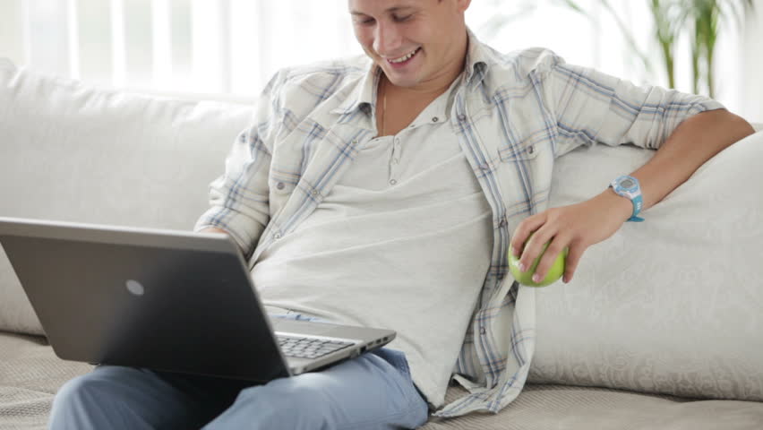 Young man sitting on sofa using laptop holding apple and smiling at camera
