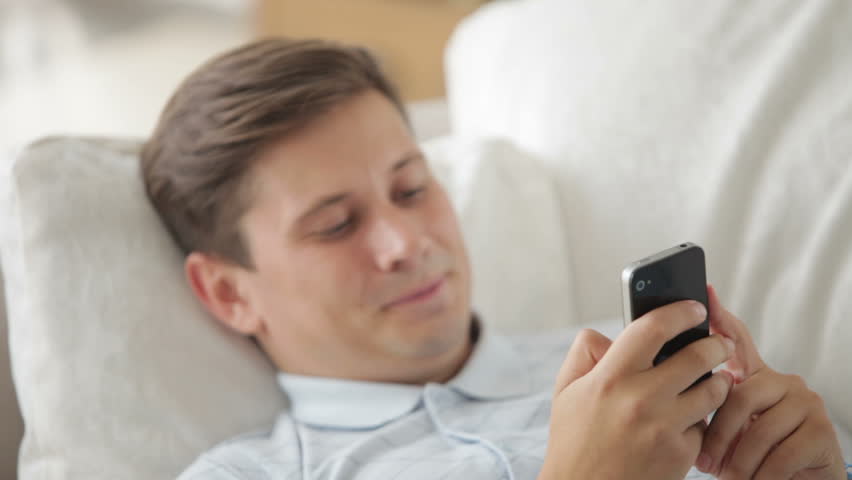Handsome young man on sofa using mobile phone and smiling at camera