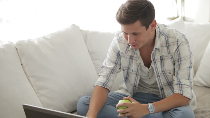 Young man sitting on sofa using laptop and eating apple