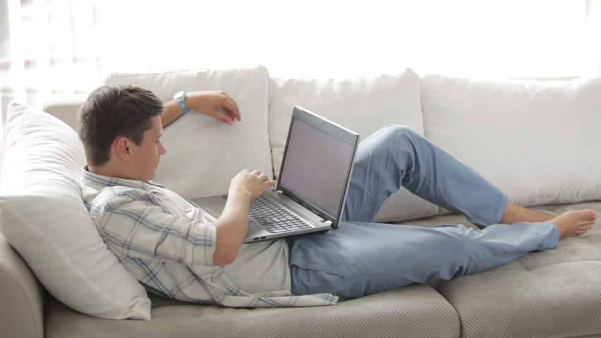 Young man lying on couch using laptop looking at camera and smiling