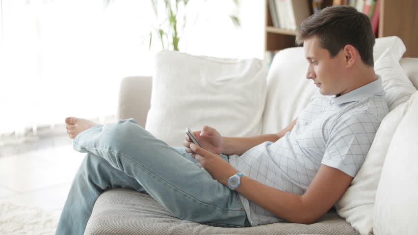 Handsome young man sitting on sofa using mobile telephone and smiling