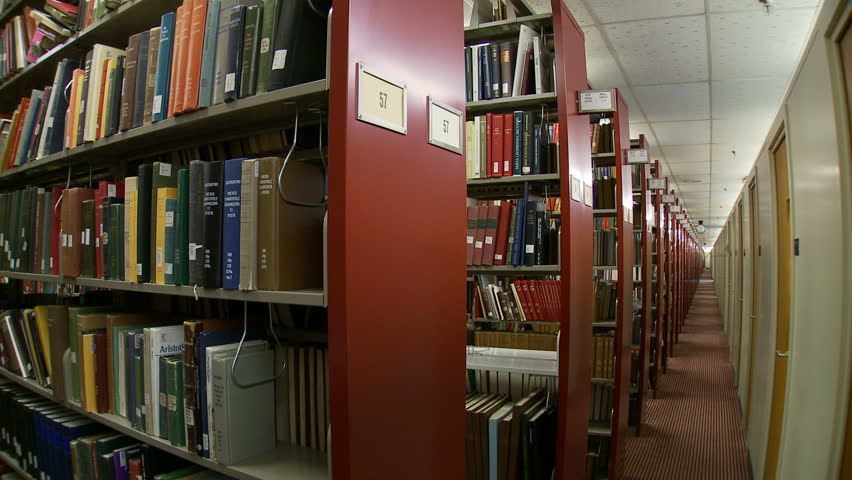 Woman walks in and out of the library stacks, down the shelves and back. 