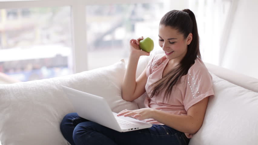 Charming young woman sitting on sofa eating apple using laptop and smiling