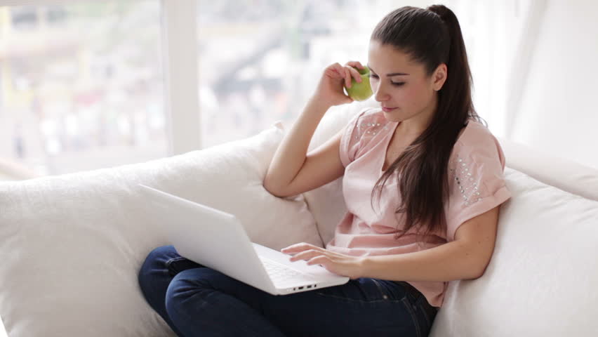 Young woman sitting on sofa with laptop holding apple looking at camera and