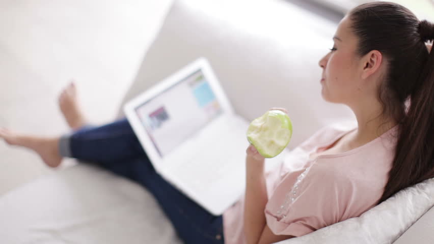 Charming young woman sitting on couch with laptop holding apple and smiling at