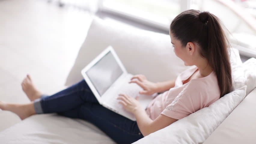 Pretty young woman relaxing on couch using laptop and looking at camera with