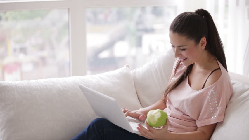 Pretty girl sitting on sofa using laptop eating apple and smiling at camera
