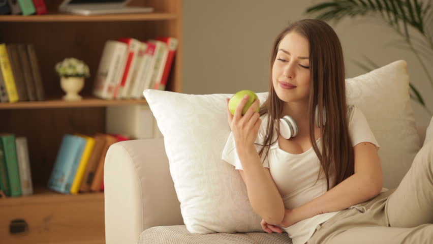 Cute girl sitting on sofa with headset eating apple and smiling at camera