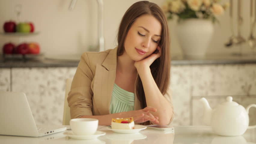 Cute girl sitting at kitchen table using mobile phone and eating cake