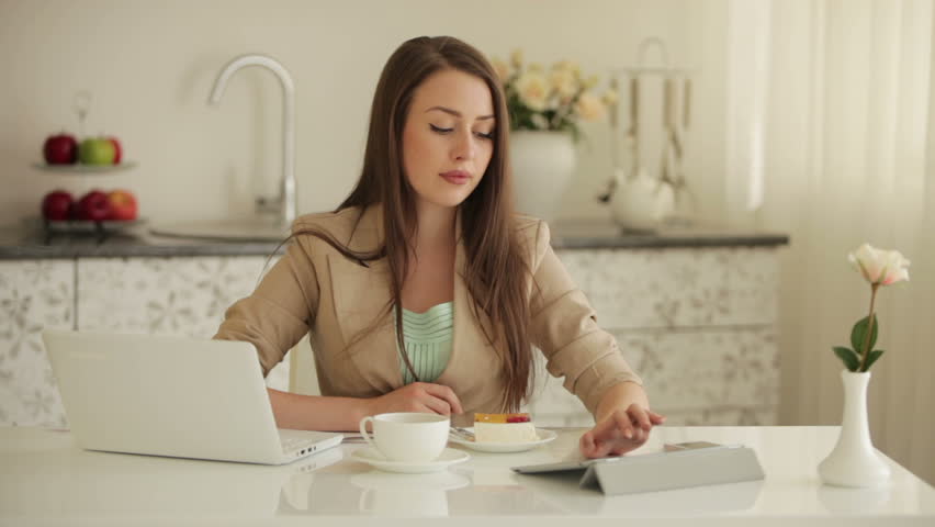 Charming girl sitting at table using laptop and touchpad drinking tea and