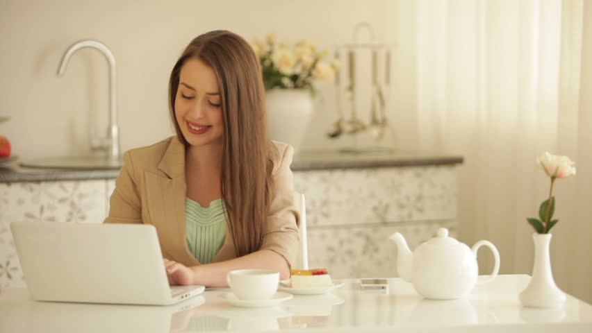 Charming young woman sitting at kitchen table with laptop eating cake drinking