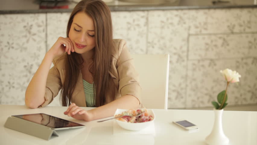 Charming young woman sitting at kitchen table with touchpad eating fruit salad