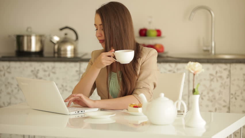 Cute young woman sitting at kitchen table with laptop drinking tea and smiling