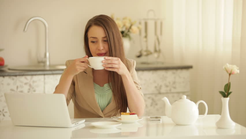 Cheerful girl sitting at table drinking tea using laptop and smiling