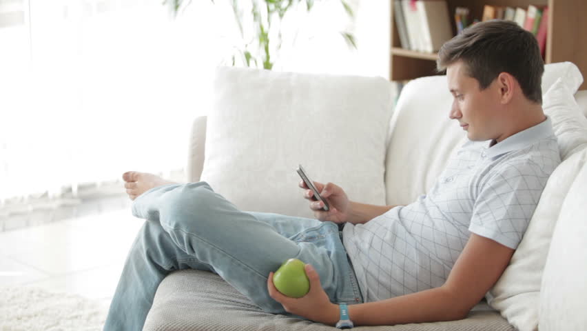 Good-looking young man sitting on sofa using cellphone eating apple and smiling