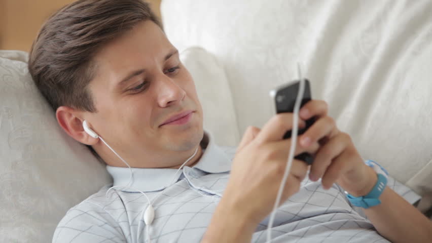 Handsome guy lying on sofa listening to music on mobile phone and smiling at