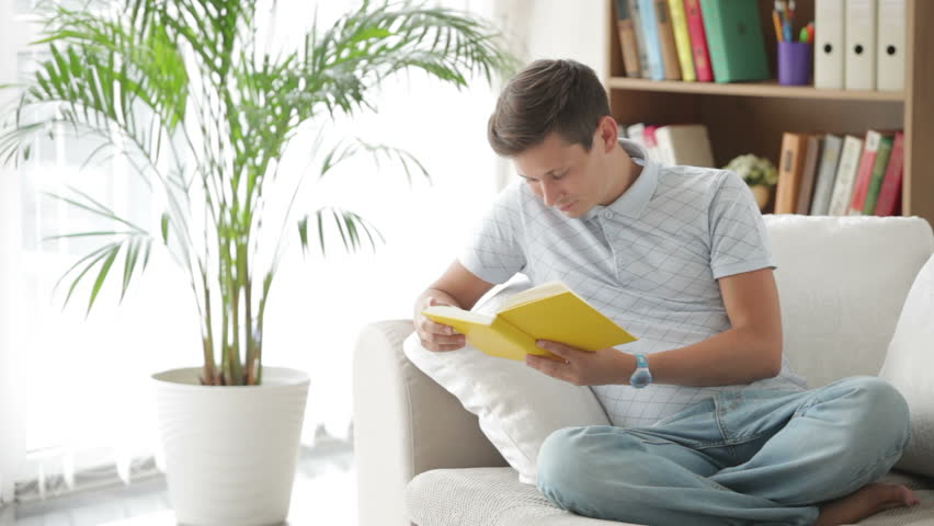 Handsome guy sitting on sofa reading book and smiling at camera