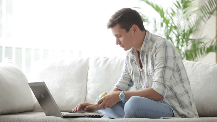 Good-looking guy sitting on sofa with laptop holding apple and looking at camera