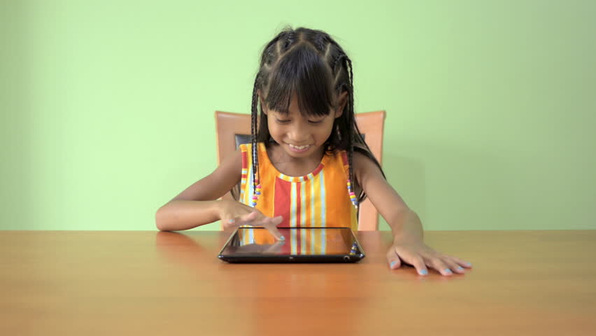 A cute young Asian girl sitting at a table playing games on tablet computer.