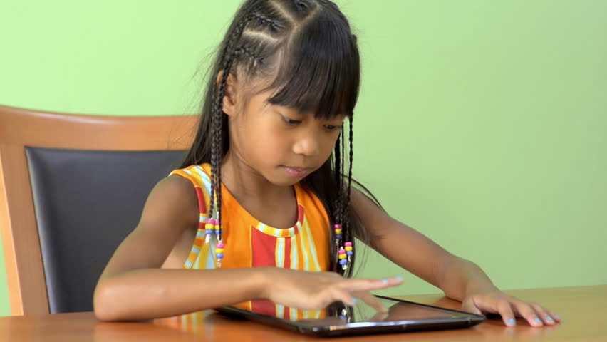 A young Asian girl sitting at a table using a tablet computer.