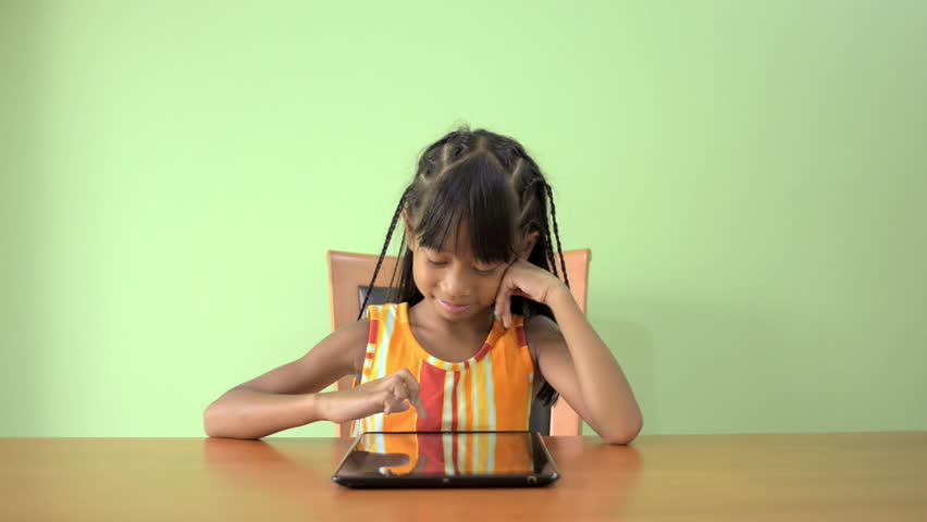 A young Asian girl sitting at a table enjoying playing on her tablet.