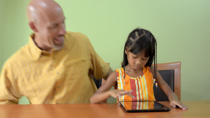 A father walks in and joins his young Asian daughter playing on a tablet