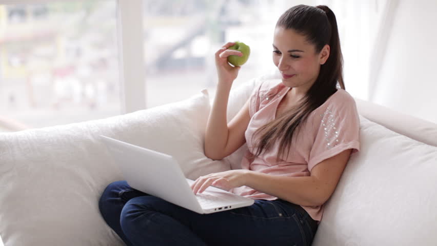 Cheerful girl sitting on sofa with laptop eating apple and smiling