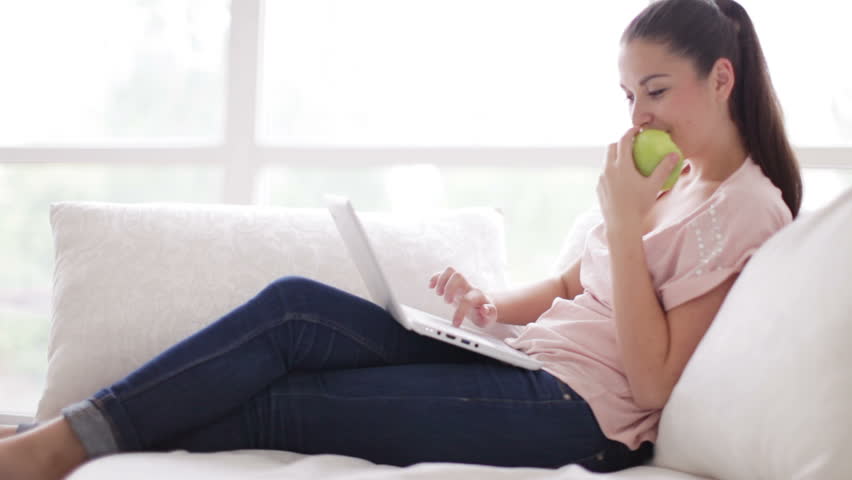 Cheerful girl sitting on couch using laptop and eating apple