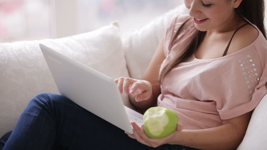 Cute brunette girl using laptop eating apple and smiling