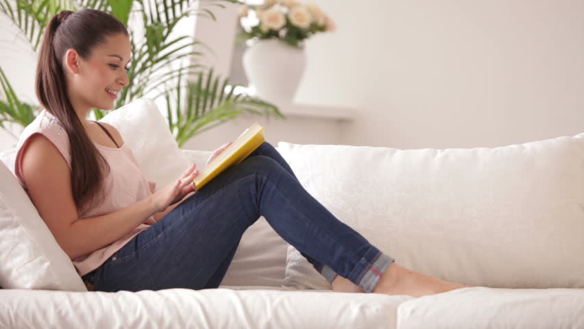 Pretty girl sitting on sofa reading book and smiling at camera