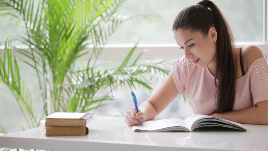 Cute girl sitting at table surrounded by books writing in notebook and smiling