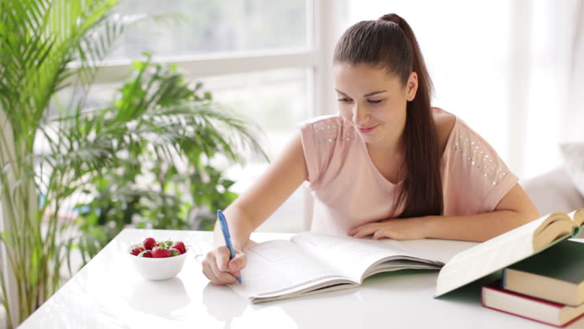 Cute girl studying at table with books and eating strawberries