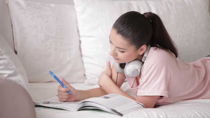 Pretty girl in headset lying on sofa writing in notebook and smiling at camera
