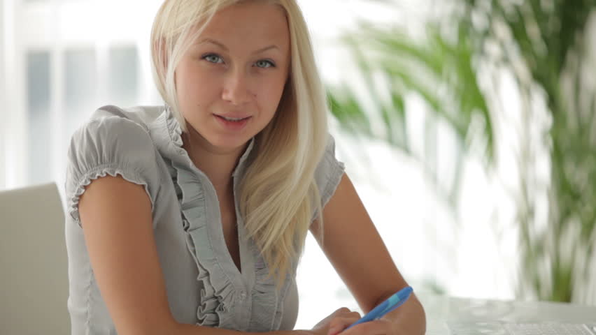 Pretty blonde girl sitting at table studying with laptop and books writing in