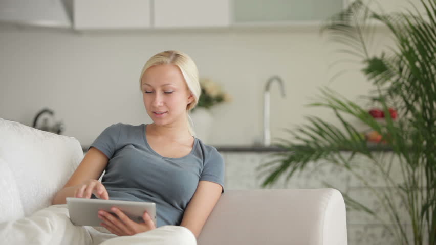 Pretty young woman sitting on sofa with touchpad and smiling at camera