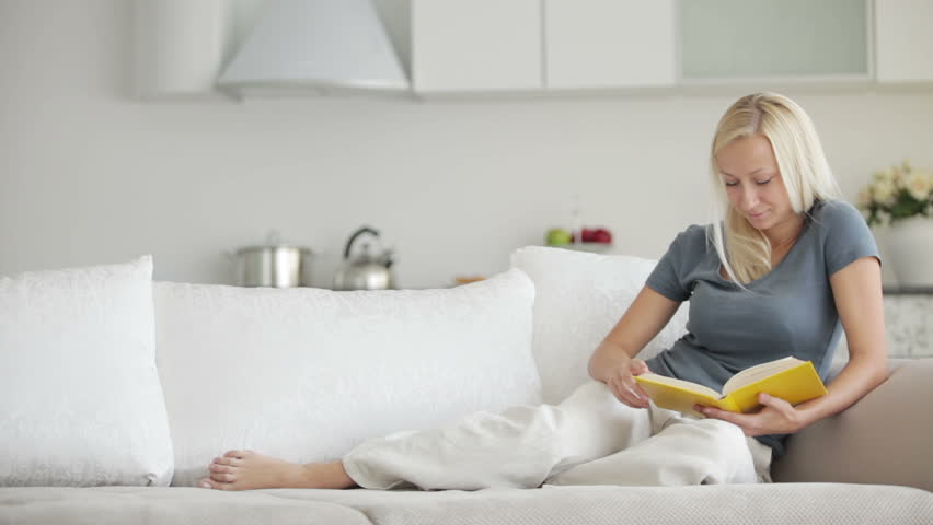 Pretty blond girl relaxing on sofa reading book and smiling at camera