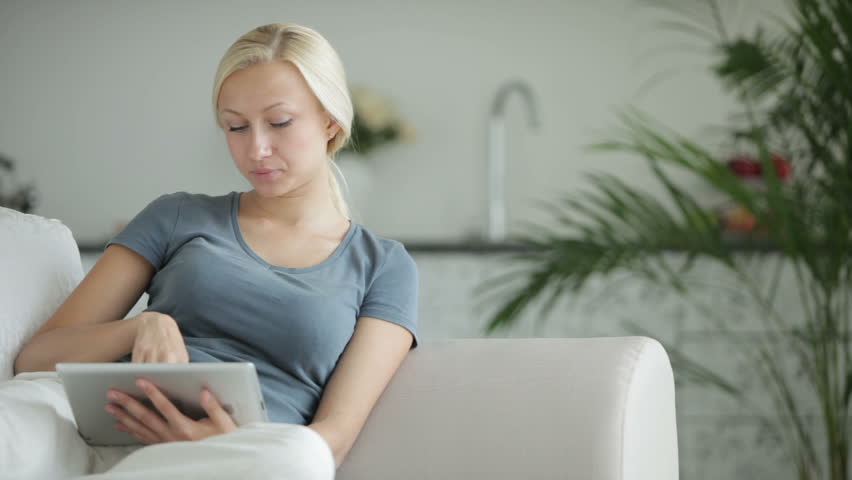 Cheerful young woman sitting on sofa using touchpad and smiling at camera