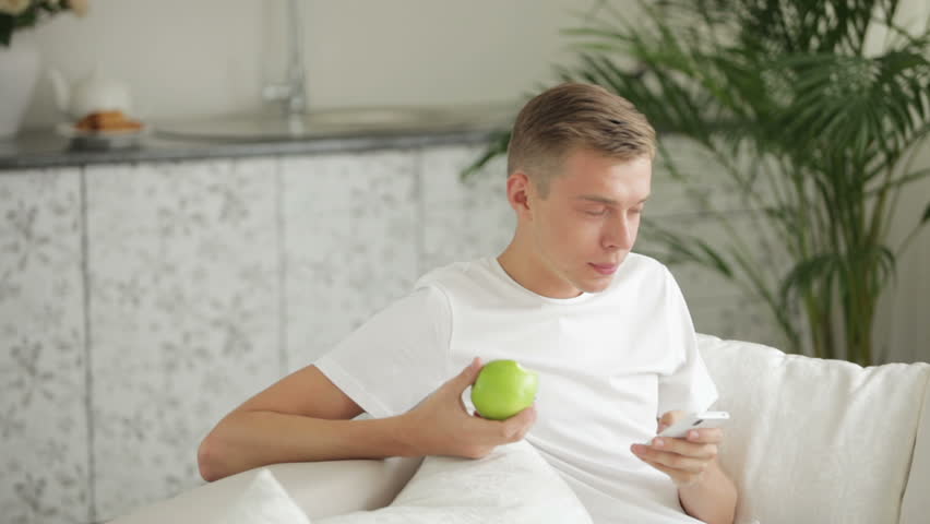 Young men sitting on sofa eating apple and using cellphone