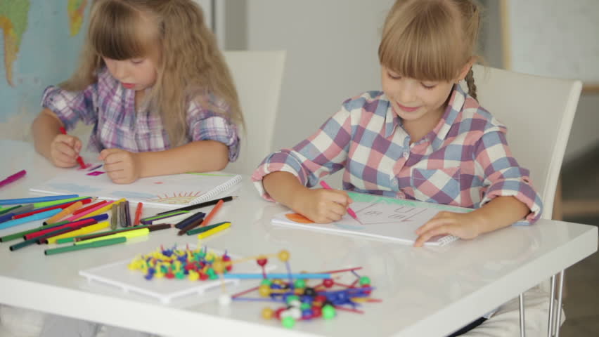Two little girls sitting at table drawing with colored pencils and smiling at