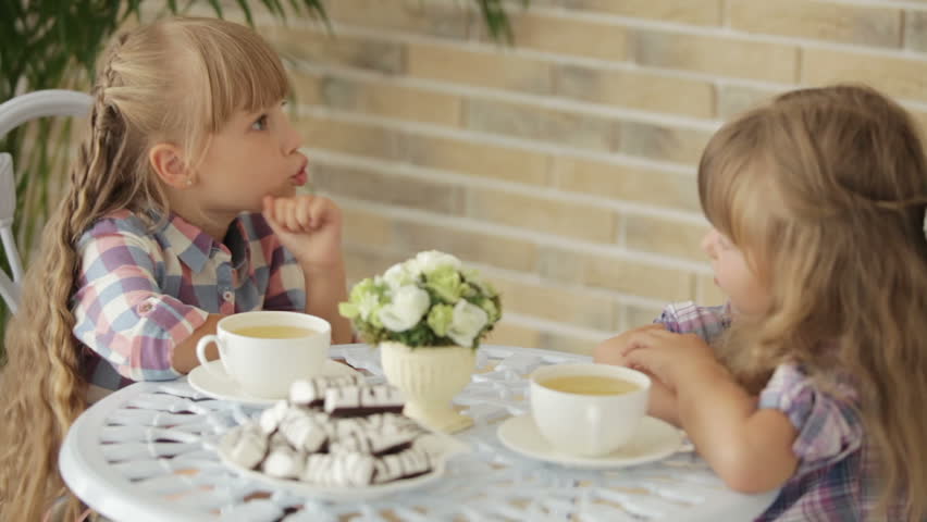 Two pretty little girls sitting at table at cafe and talking over cup of tea