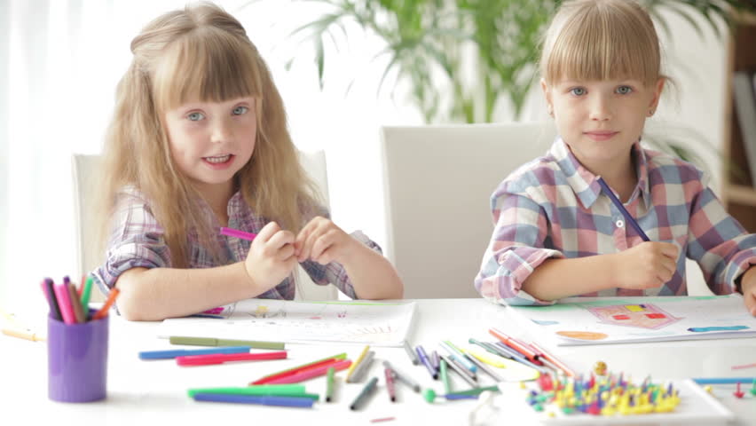 Two little girls sitting at desk drawing with colored pencils and laughing