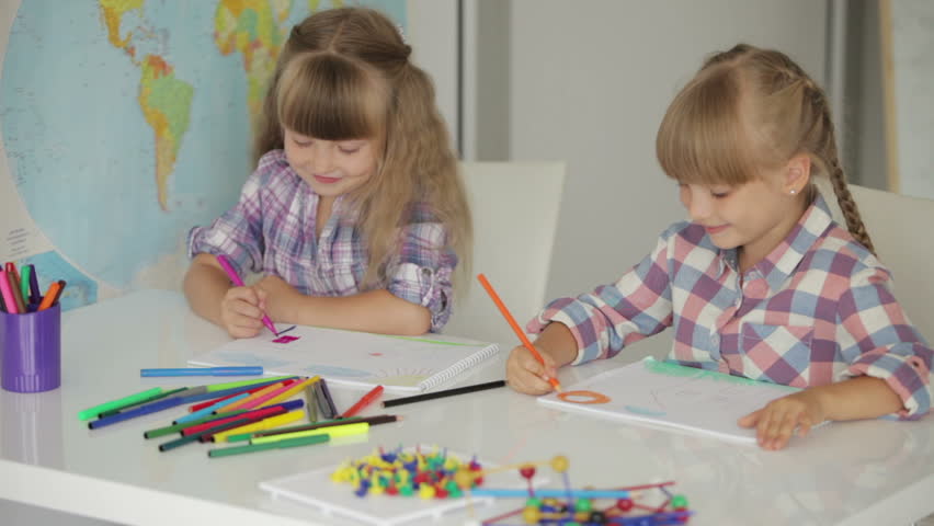 Two little girls sitting at table drawing with colored pencils and showing thumb