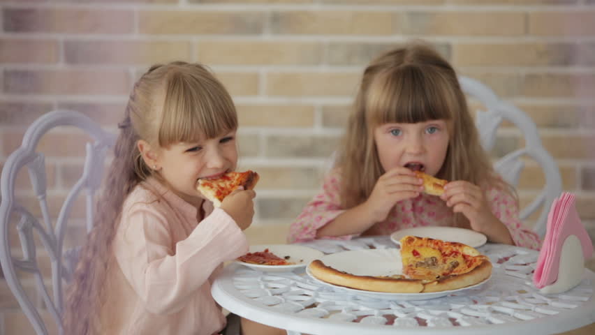 Two little girls sitting at table at cafe eating pizza and smiling